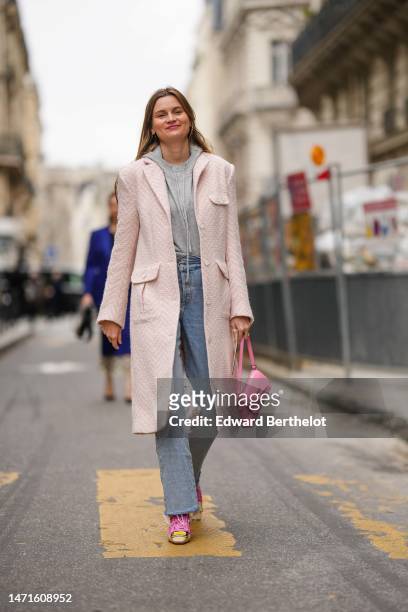 Eugenie Trochu wears gold earrings, a pale gray wool hoodie sweater, a pale pink and white braided wool long coat, blue faded denim large pants, a...