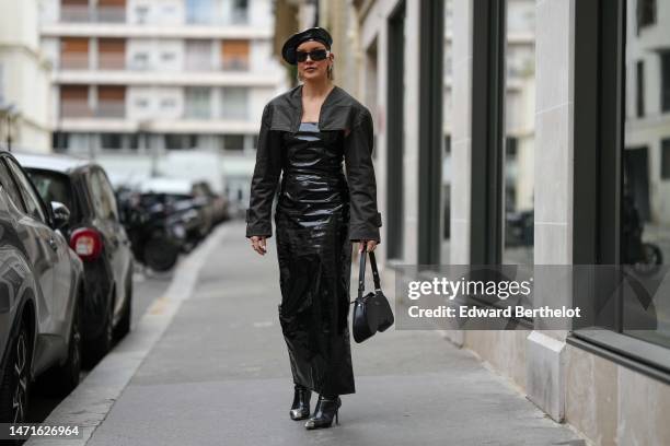 Guest wears a black shiny varnished leather beret hat, black sunglasses from Chanel, silver earrings, a black faded denim cropped jacket, a black...