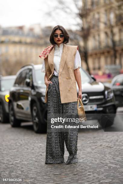 Guest wears black and gold sunglasses, a white high neck buttoned blouse short sleeves shirt, a beige linen sleeveless blazer gilet, a beige wicker...