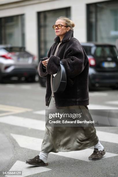 Guest wears black and yellow sunglasses, a black sheep fur oversized coat, a khaki gray silk long dress, a black shiny leather handbag, white socks,...