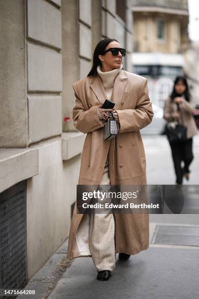 Guest wears black sunglasses, a beige ribbed wool turtleneck pullover, a beige shiny leather long coat, beige wool pants, a black suede with...