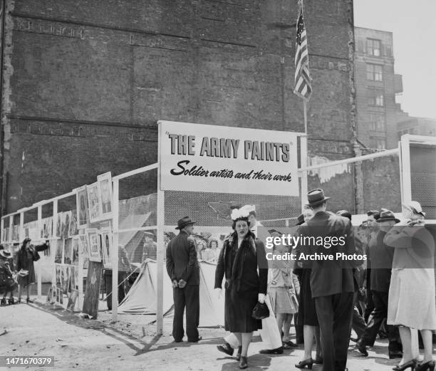 Group of people attend an open-air exhibition displayed beneath a sign reading 'The Army Paints - Soldier artists and their work' and an American...