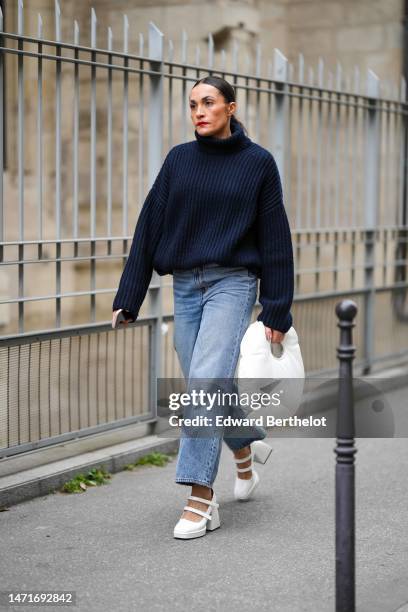 Guest wears a navy blue ribbed wool turtleneck pullover, blue denim large pants, a white shiny leather puffy handbag, white shiny leather belted...