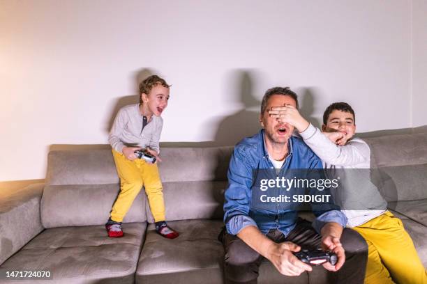 boy covering eyes to his father, so his brother can win, while playing video games on playstation against father - being the best dad he can be stock pictures, royalty-free photos & images