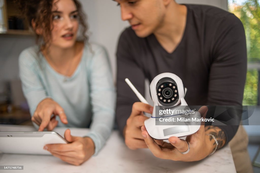 A young couple, a man and a woman, are setting up a security camera for home surveillance in their apartment