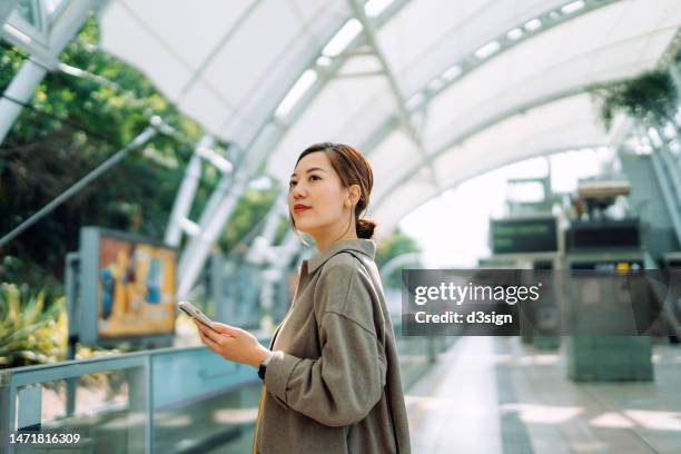 confident young asian businesswoman using smartphone while waiting for the train in subway station. commuting to work. travelling on public transportation in the city. technology in everyday life. business on the go - day in the life stock pictures, royalty-free photos & images