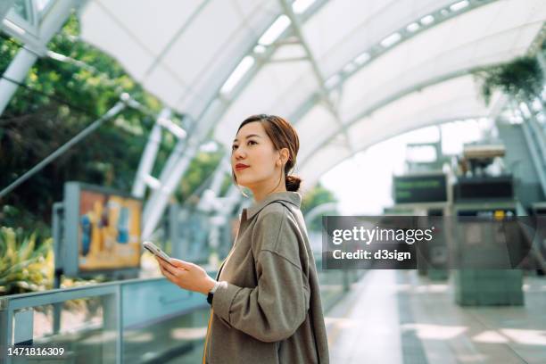confident young asian businesswoman using smartphone while waiting for the train in subway station. commuting to work. travelling on public transportation in the city. technology in everyday life. business on the go - um dia na vida de imagens e fotografias de stock