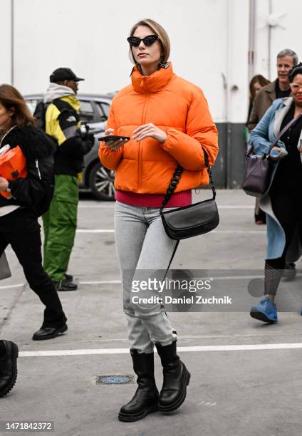 Guest is seen wearing an orange puff jacket, gray jeans, black boots, black bag and black and gold sunglasses outside the Rokh show during Paris...