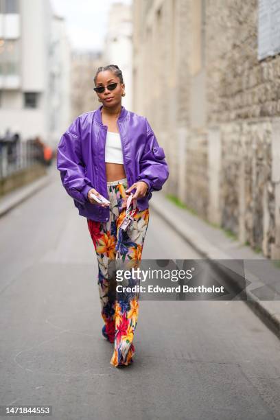 Ellie Delphine wears black vintage sunglasses, a white cropped tank-top, a purple shiny leather oversized bomber coat, high waist beige with navy...