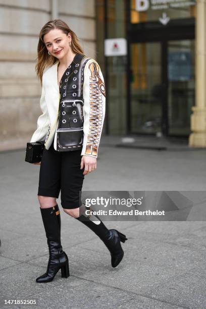Chloe Grace Moretz wears gold and diamonds earrings from Louis Vuitton, a white shiny leather with gold and black print pattern blazer jacket from...