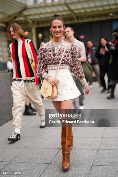 Alicia Vikander wears white pearl and gold earrings, a white with red and black flower print pattern pleated / long puffy sleeves blouse, a white...