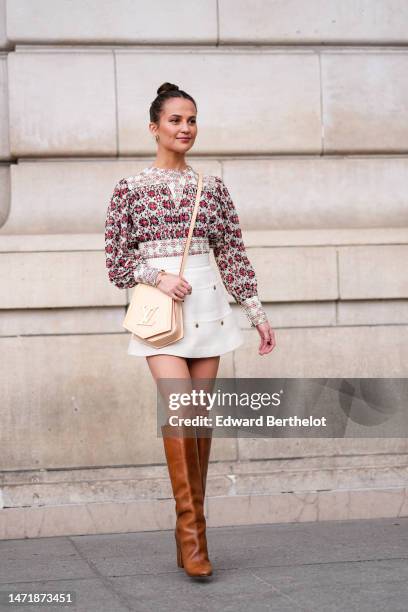 Alicia Vikander wears white pearl and gold earrings, a white with red and black flower print pattern pleated / long puffy sleeves blouse, a white...