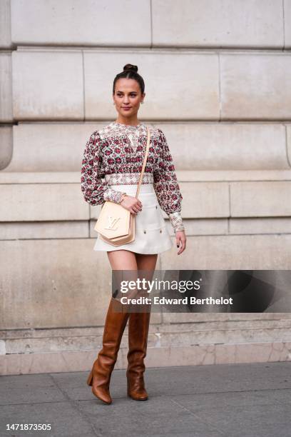 Alicia Vikander wears white pearl and gold earrings, a white with red and black flower print pattern pleated / long puffy sleeves blouse, a white...
