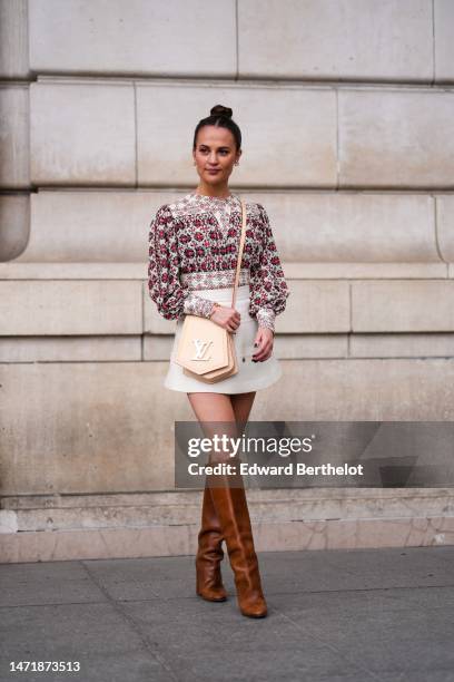 Alicia Vikander wears white pearl and gold earrings, a white with red and black flower print pattern pleated / long puffy sleeves blouse, a white...