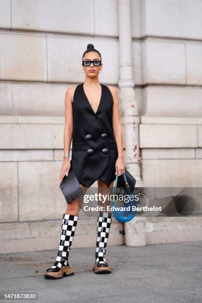 Jessica Aidi wears black and blue squared sunglasses, diamond earrings, a black V-neck / sleeveless / large leather buttons asymmetric blazer short...