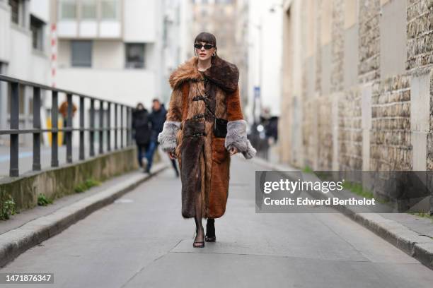 Mary Leest wears black sunglasses, silver earrings, a black t-shirt, a brown / camel / gray embroidered yoke fur long coat, a black shiny leather...