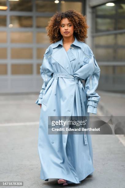 Paola Locatelli wears a pale blue shiny leather belted and buttoned long coat, a pale blue shiny leather pleated / accordion short dress, pale pink...