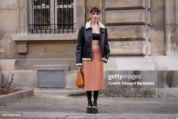 Mary Leest wears gold earrings, a black shoulder-off / cropped top, a black shiny leather with beige sheep collar zipper jacket, a camel high waist...