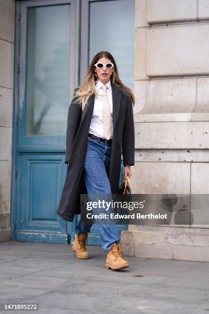 Guest wears white sunglasses, white pearls earrings, a white shirt, a beige and gold checkered print pattern silk tie, a dark gray long coat, a brown...