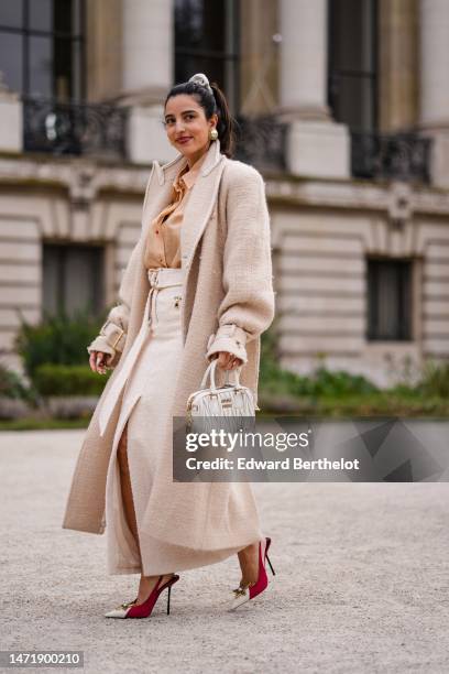 Guest wears a white ribbed velvet hair elastic, gold and white pearls large earrings, a pale orange shirt, a beige tweed long oversized coat, a white...