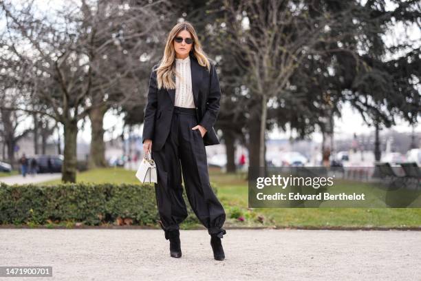 Guest wears black sunglasses, a white lace print pattern high neck blouse, a black blazer jacket, black puffy suit pants, a white shiny leather...