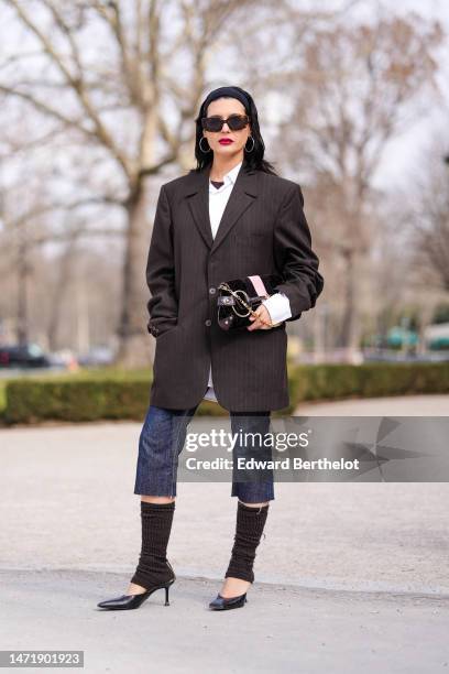 Guest wears brown marble sunglasses, a black large headband, silver earrings, a black t-shirt, a white shirt, a dark brown oversized blazer jacket, a...