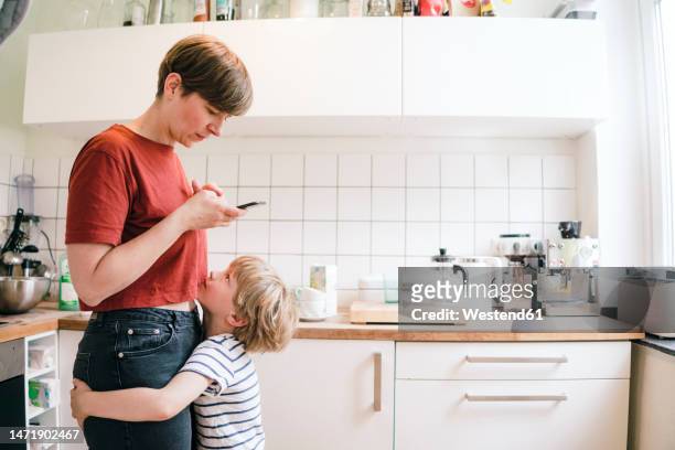 boy embracing mother using smart phone standing in kitchen - child mobile phone stock-fotos und bilder