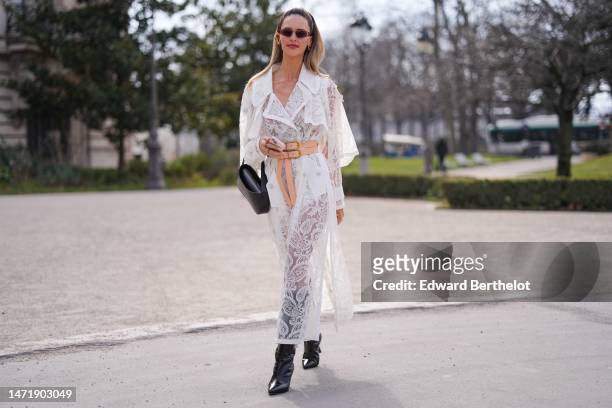Guest wears burgundy sunglasses, a white lace print pattern cut-out details long dress, a beige shiny leather large belt, a black shiny leather tube...