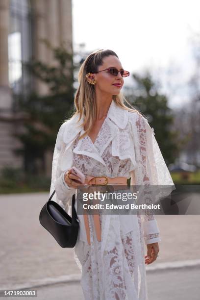 Guest wears burgundy sunglasses, a white lace print pattern cut-out details long dress, a beige shiny leather large belt, a black shiny leather tube...