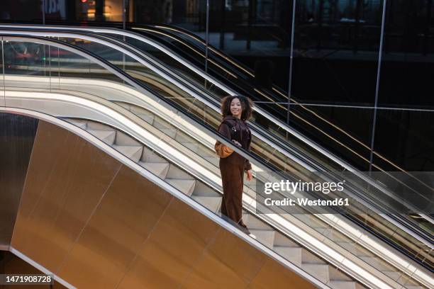 smiling young woman moving down escalator - three quarter length stock pictures, royalty-free photos & images