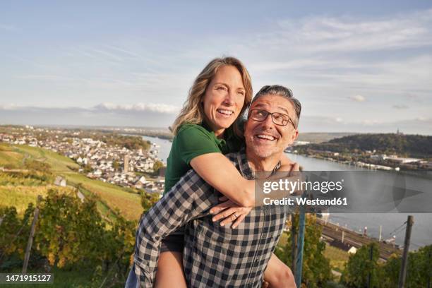 happy mature man giving piggy back ride to woman on sunny day - rheingau stock pictures, royalty-free photos & images