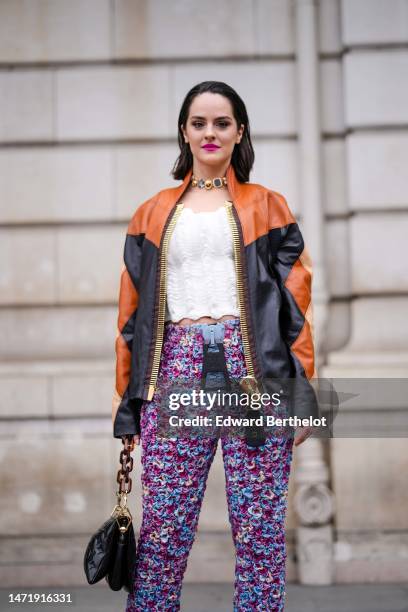 Guest wears a gold with black stones large necklace, a white wavy ruffled shoulder-off top, a black / orange / beige shiny leather print pattern...