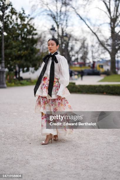 Guest wears gold earrings, a black silk knot long tie, a white oversized / puffy sleeves blouse, a white with orange / red / pink flower print...