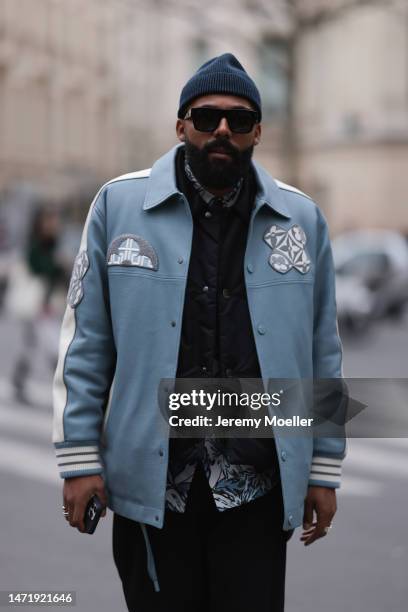 Jean-Claude wears black sunglasses, a blue hat, a Louis Vuitton blue leather jacket with white details, a black vest, a shirt with flower pattern, a...