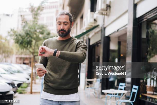 man holding smart phone checking time on wristwatch - puntualidad fotografías e imágenes de stock