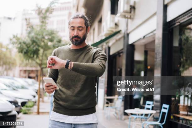 man holding smart phone checking time on wristwatch - checking watch stock pictures, royalty-free photos & images