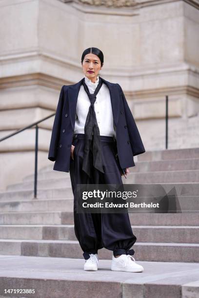 Guest wears a white ruffled high neck blouse, a black silk tie, a black buttoned coat, black puffy pants, white shiny leather sneakers , outside...
