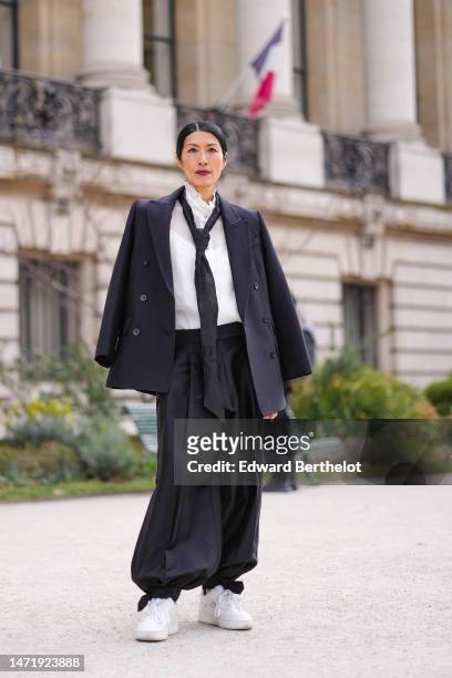 Guest wears a white ruffled high neck blouse, a black silk tie, a black buttoned coat, black puffy pants, white shiny leather sneakers , outside...