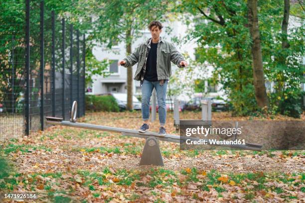 young man standing and balancing on seesaw in park - seesaw stock pictures, royalty-free photos & images