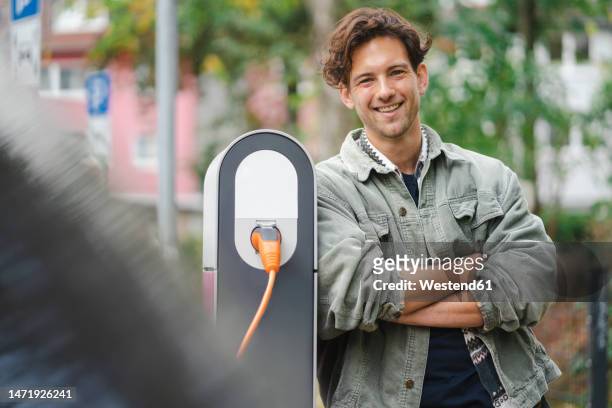 happy young man leaning on electric vehicle charger - stromtankstelle stock-fotos und bilder