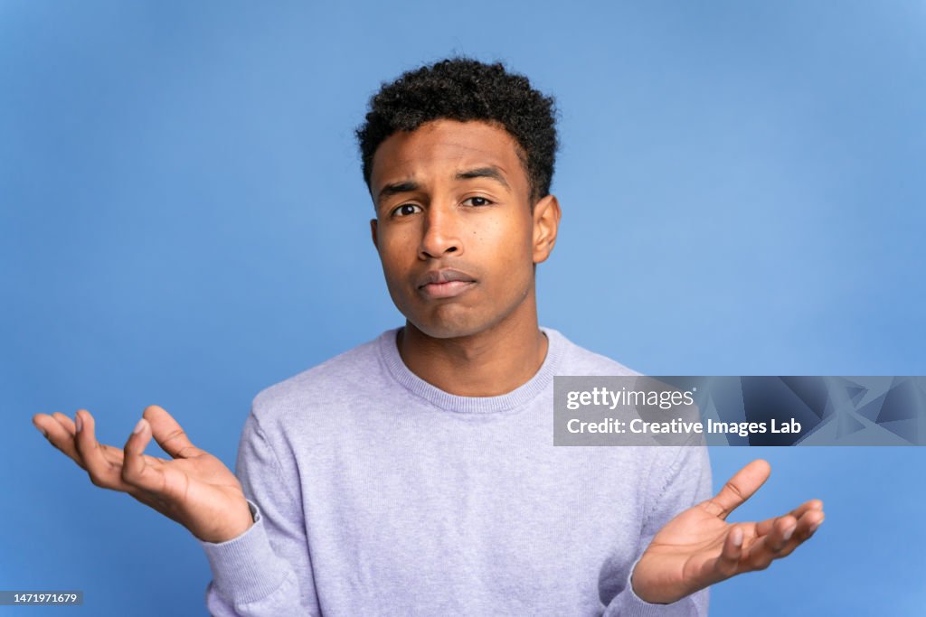 Young African American male in casual clothes showing doubt gesture and looking at camera while standing against blue background