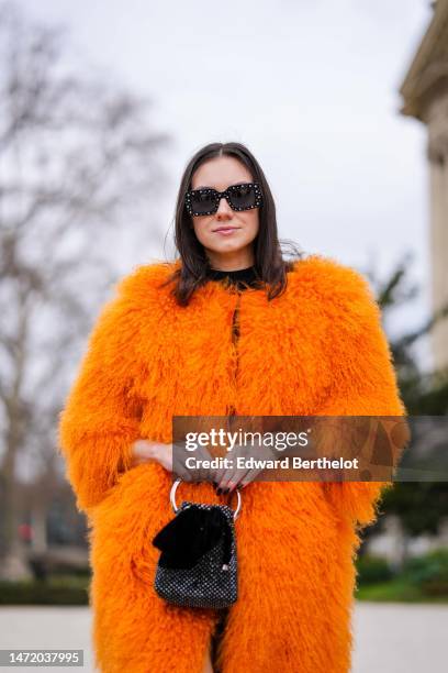 Guest wears black with white polka dots print pattern sunglasses, a black turtleneck pullover, a neon orange fluffy fur oversized long coat, a black...
