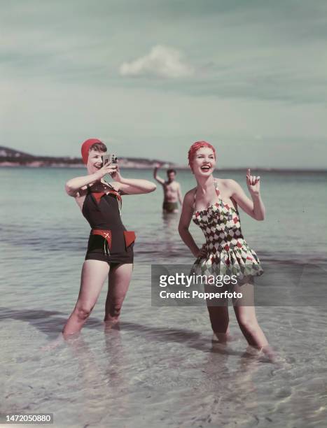 Vacation scene of two female fashion models posed paddling in the sea wearing swimsuits, one suit is black with contrasting red pockets, and the...
