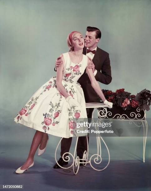 Posed studio portrait of a young couple, the woman wears a white party dress in a pink rose print with a full skirt, the man wears a dark brown suit...