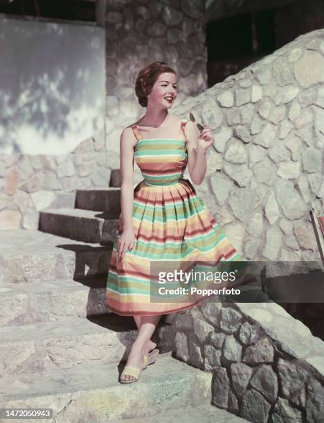 Vacation scene of a female fashion model posed wearing a strappy sun dress in broad striped fabric, the dress features a fitted bodice and a full...