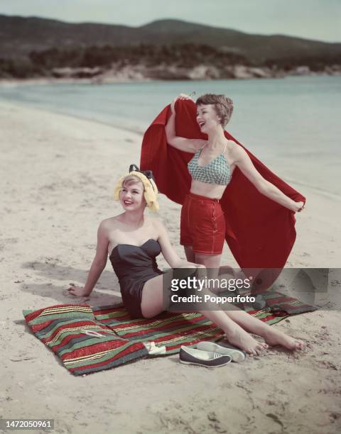 Vacation scene of two female fashion models posed on a sandy beach, one wears a strapless black swimsuit, the other wears a green and white print...