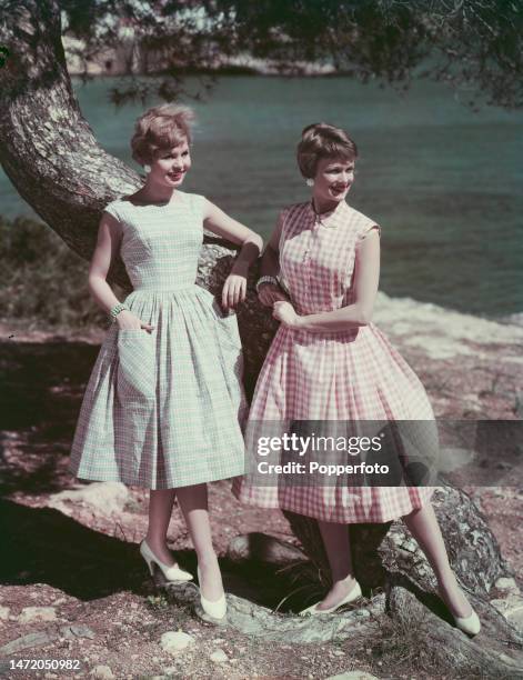 Vacation scene of two female fashion models posed wearing summer dresses beside a tree in a seaside setting, one dress is in a green check fabric...