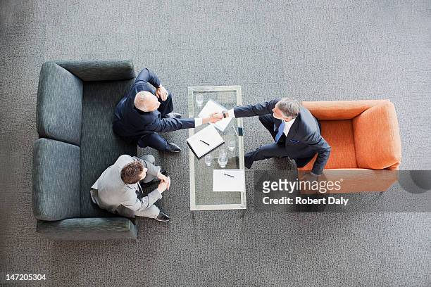 businessmen shaking hands across coffee table in office lobby - business agreement stock pictures, royalty-free photos & images