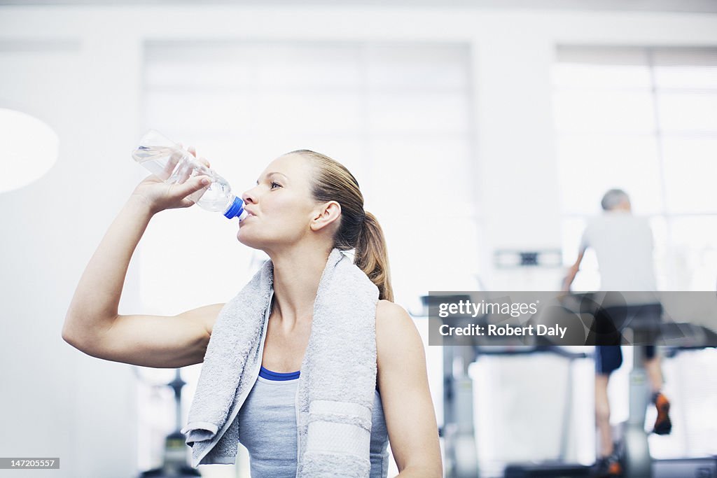 Woman with towel around neck drinking water in gymnasium