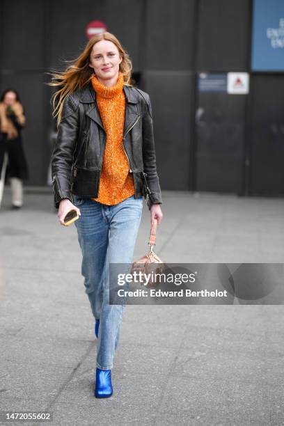 Guest wears an orange wool turtleneck pullover, a black shiny leather zipper jacket, blue faded denim pants, a pale pink satin handbag from Fendi,...